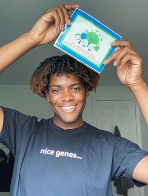 Brandon Solis is wearing a black "nice genes..." T shirt and holding up a card with Addgene mascots, Blugene and Aavery, on it. The card says "Welcome to Addgene! We are so excited to have you"