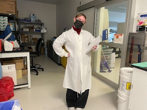 Woman in a white lab coat holding a clipboard and smiling in a lab. 