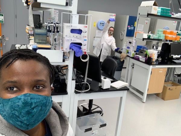 Two Addgenies in the lab social distancing wearing masks. One Addgenie is taking the selfie, the other Addgenie is in the background