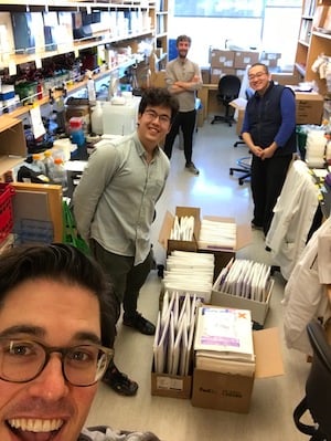 Four scientists in the lab standing between lab benches with boxes of packages containing plasmids ready to ship out