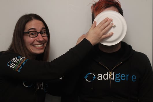 A young, smiling woman wearing an Addgene 10 year anniversary smashes a pie into the face of a man wearing the same shirt.