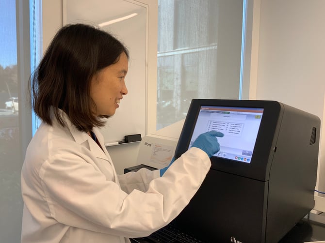 A woman in PPE points to the touchscreen of an NGS sequencer.