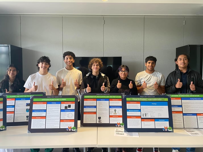 Seven interns standing behind their posters and smiling with thumbs-up. 